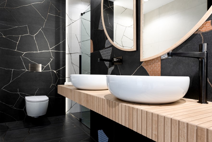Łazienka z ciemnymi geometrycznymi płytkami z elementami słota i blatem z płytek drewnopodobnych.