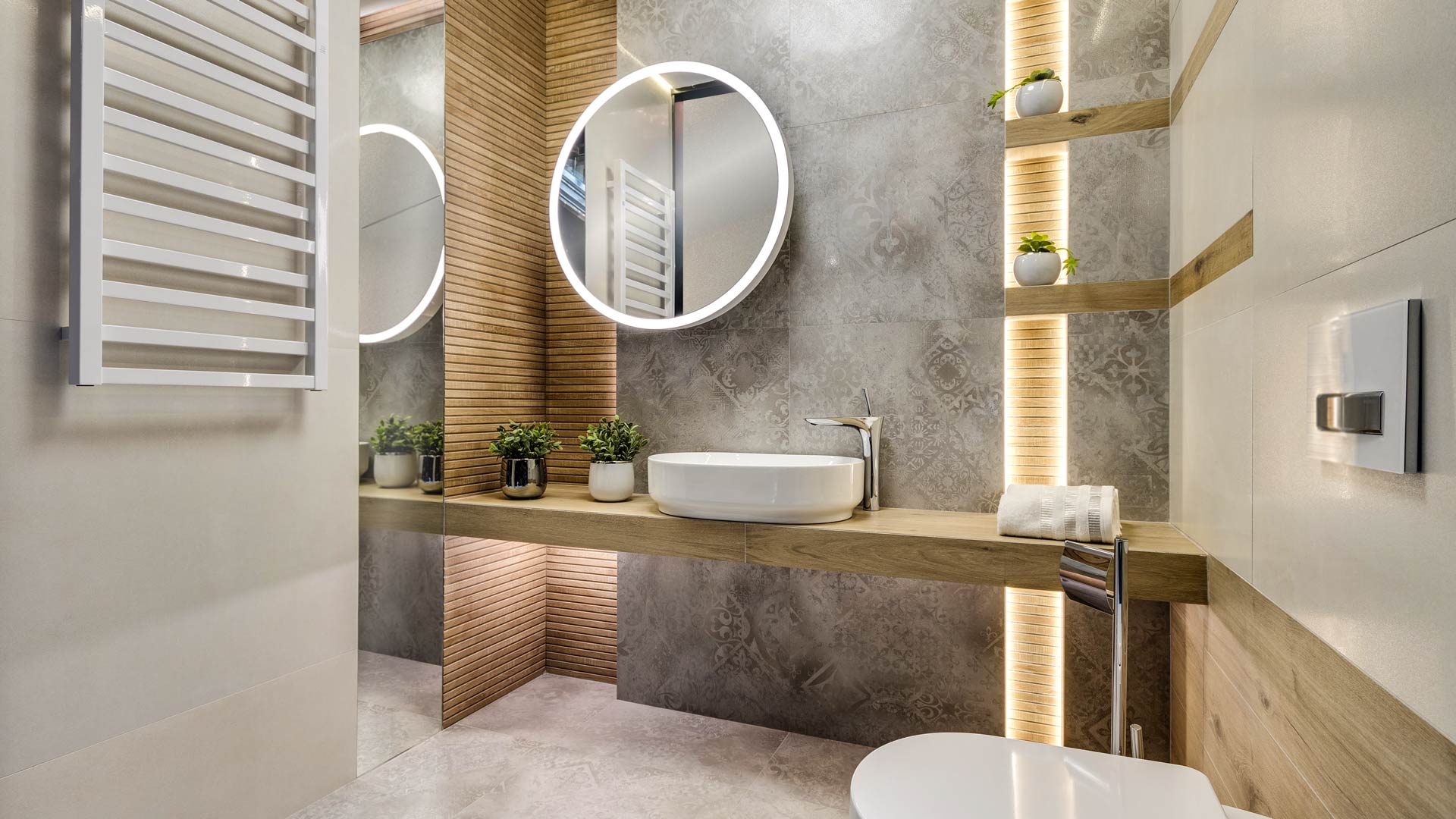 ciepła łazienka z długim drewnianym blatem, delikatnie wzorzystą szarą płytką, okrągłym podświetlanym lustrem, małymi doniczkami i pionowymi lamelami z LED‑ami